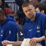 New Golden State Warriors forward Kristaps Porzingis (with towel) watches the action against the Boston Celtics from the bench during the first quarter at Chase Center