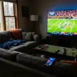 Empty living room couch facing TV showing NFL game with streaming apps on phone, representing fan accessibility debate