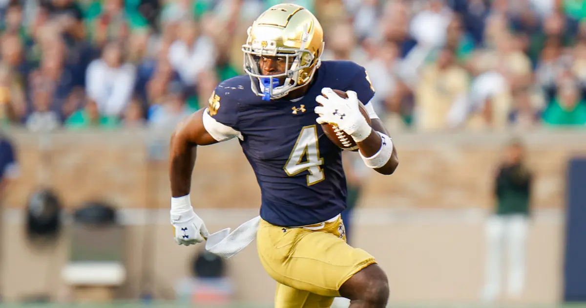 Notre Dame football player in blue jersey running with the ball during a game.