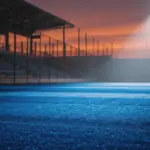 Floodlit stadium pitch at dusk with dramatic shadows, no players visible, cool blue lighting across empty turf.