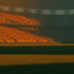 Floodlit stadium at golden-hour kick-off with packed stands, dramatic shadows on pristine pitch, no players visible.