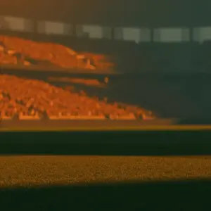 Floodlit stadium at golden-hour kick-off with packed stands, dramatic shadows on pristine pitch, no players visible.
