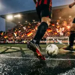 Floodlit stadium at evening with German national colours in crowd, football boot on wet pitch, dramatic shadows and net.