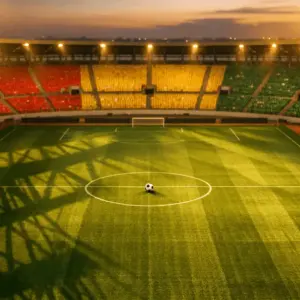 Aerial view of floodlit stadium pitch with Ghana flag colours in stands and ball on centre spot at dusk.