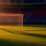 Empty stadium pitch at golden-hour kick-off, warm lighting and dramatic shadows, red and blue sectioned stands