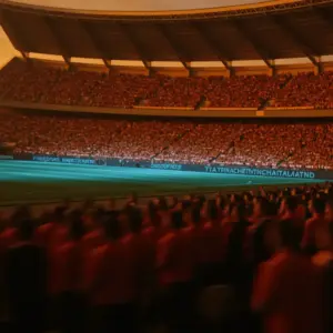 Floodlit stadium with packed orange and red crowd at golden-hour dusk, low angle, dramatic shadow and light contrast.