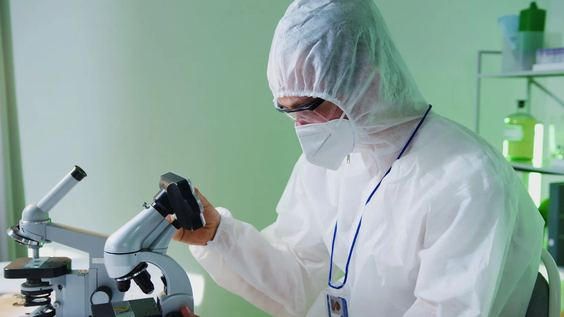 Laboratory scientist in protective gear examining samples with a microscope.