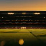 Floodlit football stadium at dusk with packed stands, dramatic shadow patterns, and golden-hour pitch lighting before kickoff.