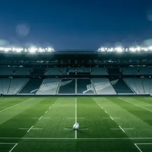 Floodlit stadium pitch at dusk with All Blacks national colours in stands, centre-spot football, dramatic shadow geometry.