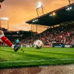 Football player in mid-kick on floodlit stadium pitch with red and white crowd in background during dusk.