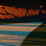Golden-hour floodlit stadium pitch with geometric shadows and packed stands, shallow depth of field, cinematic lighting.