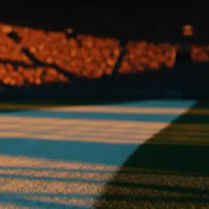 Golden-hour floodlit stadium pitch with geometric shadows and packed stands, shallow depth of field, cinematic lighting.