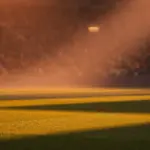 Floodlit stadium pitch at dusk, geometric shadow patterns, packed stands in bokeh, yellow-green and blue-red colour tones.