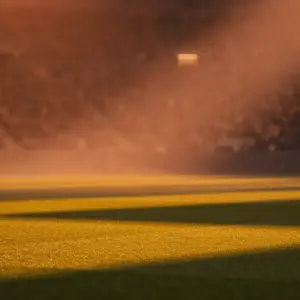 Floodlit stadium pitch at dusk, geometric shadow patterns, packed stands in bokeh, yellow-green and blue-red colour tones.