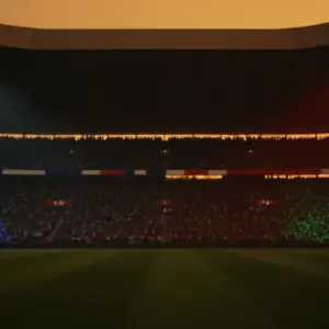 Floodlit stadium at dusk with dramatic shadows, blue and red colour grading, packed stands in silhouette.