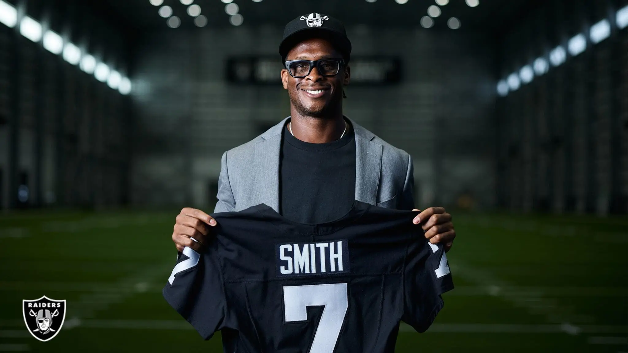 Geno Smith wearing a Raiders hat and blazer, holding up a black Las Vegas Raiders jersey with SMITH 7 on the back.