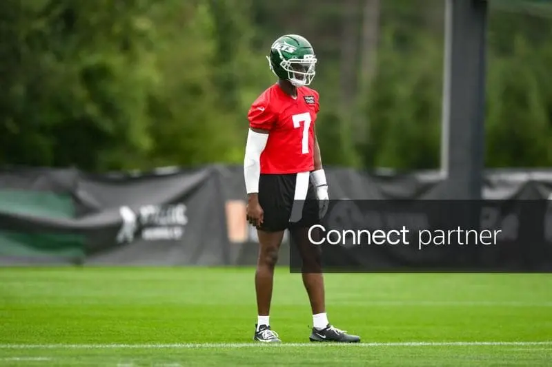 New York Jets player wearing a red jersey and number 7 during a practice session on a grass field.