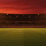 Empty floodlit stadium at dusk with dramatic shadows and golden-hour lighting creating atmospheric match preview mood.