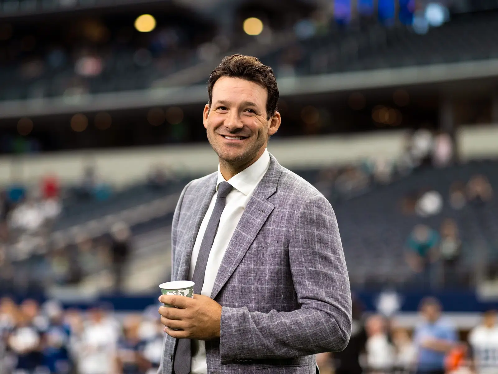Tony Romo smiling and holding a cup while wearing a grey blazer and tie at a football stadium