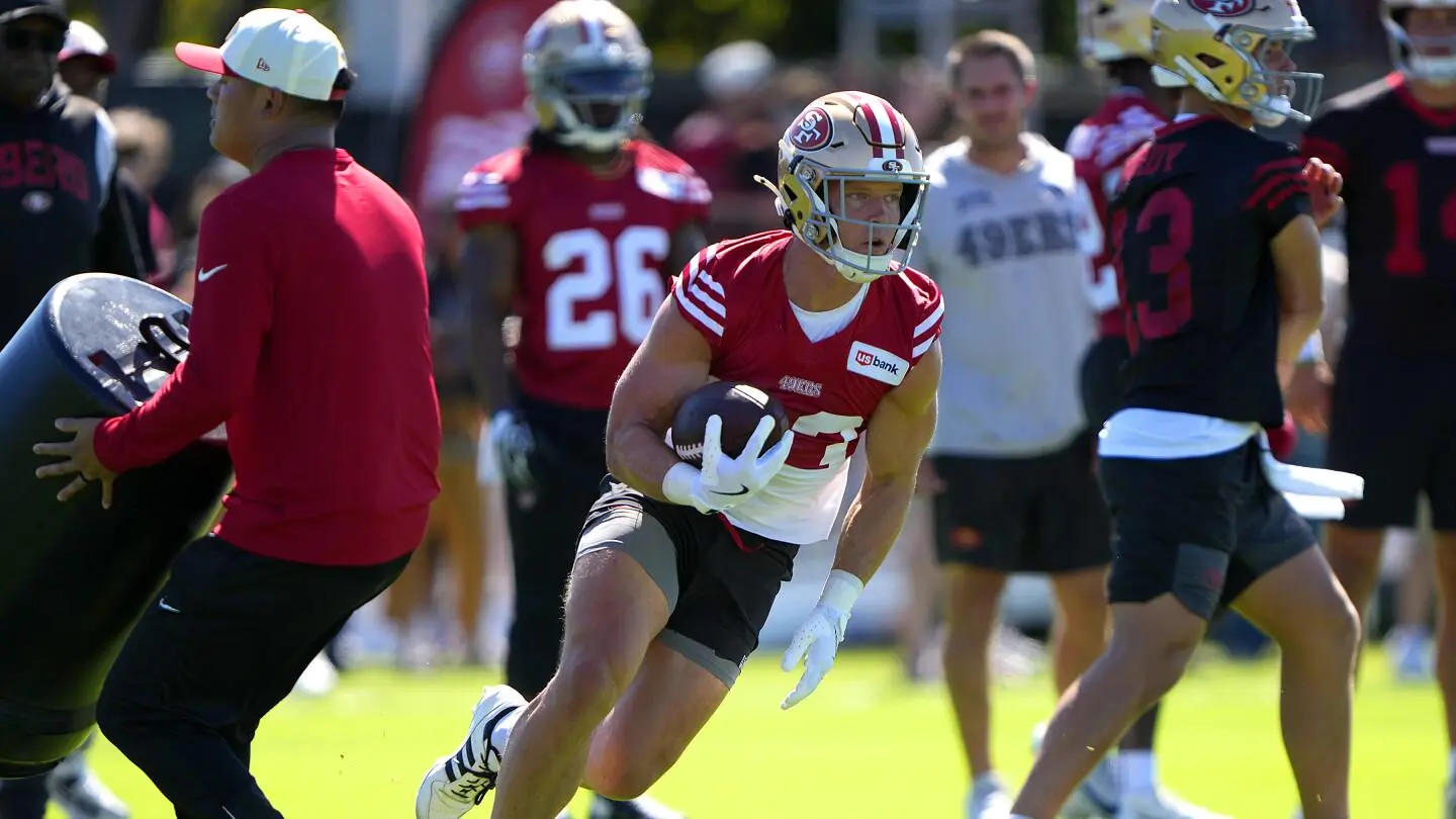 Christian McCaffrey wearing a San Francisco 49ers practice jersey and helmet running with a football