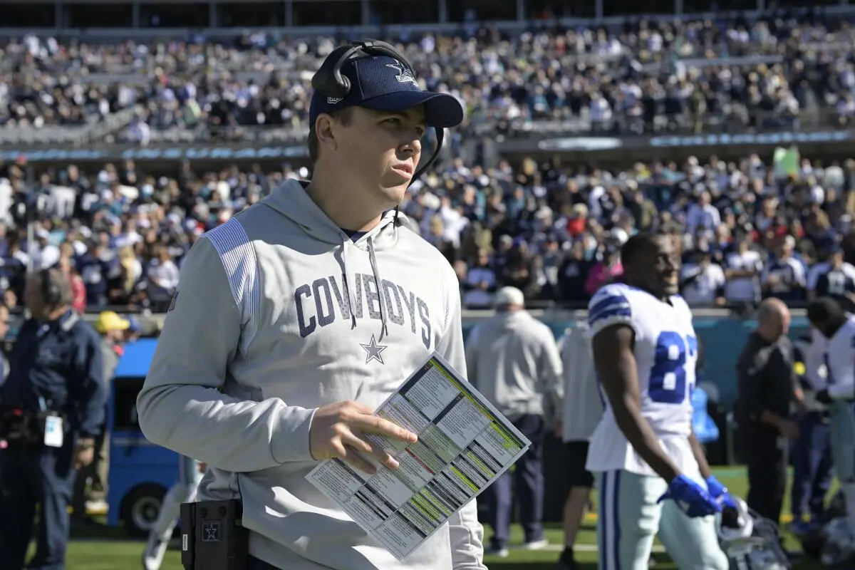Kellen Moore wearing a Dallas Cowboys hoodie and headset on the sideline holding a play sheet