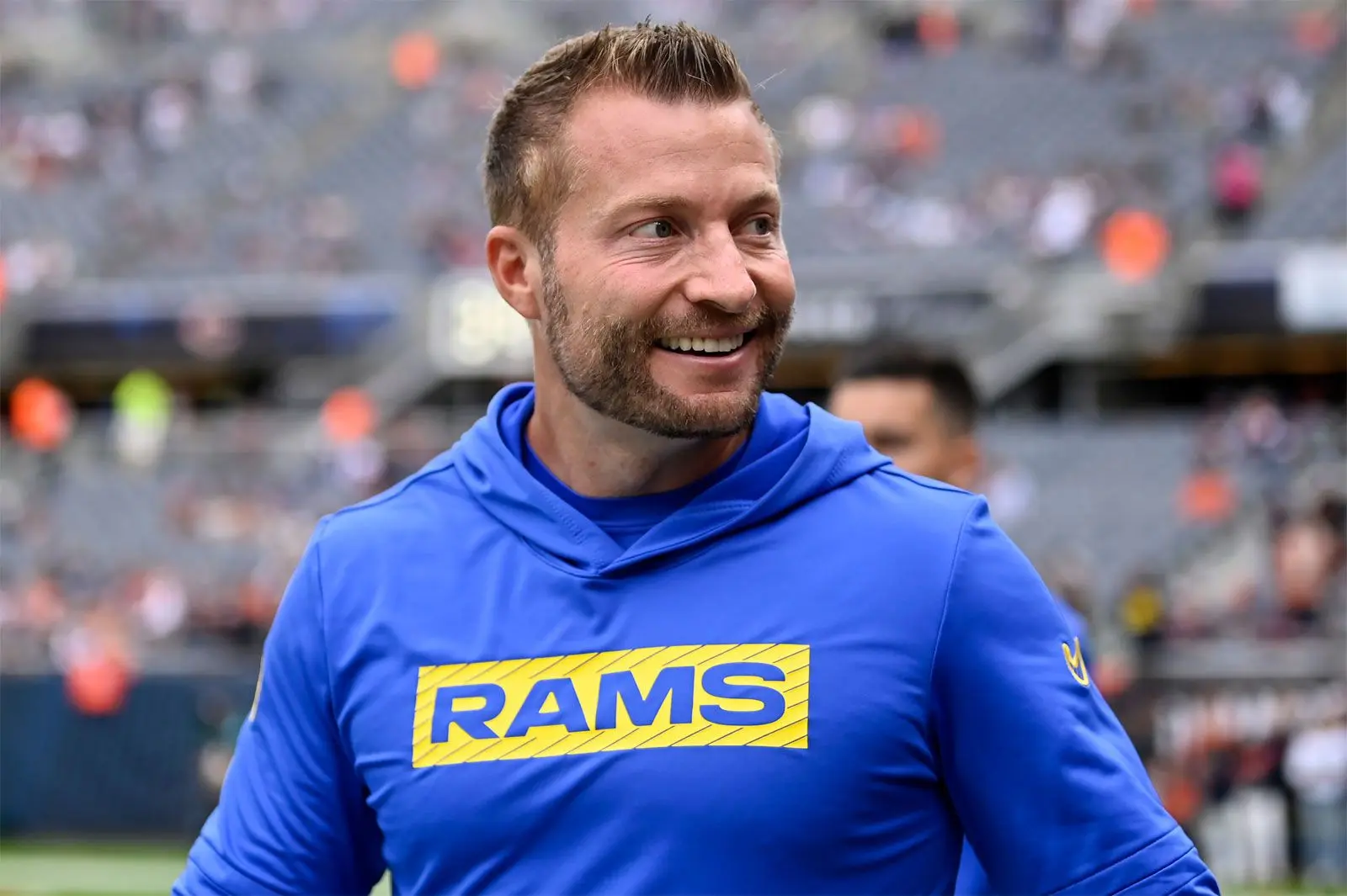 Sean McVay smiling while wearing a blue Los Angeles Rams hooded sweatshirt on a football field.